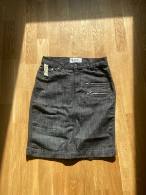 Svarta Pudel jeanskjol med dragkedjor - Snygg svart jeanskjol från Pudel med två dekorativa dragkedjor framtill och bälteshällor. Kjolen har en klassisk rak passform och är perfekt för dig som gillar en stilren look med edge. Materialet är denim och kjolen har normal midja.