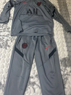 Grå PSG träningsset från Nike - Säljer ett grått träningsset från Nike med PSG-logga. Setet består av långärmad tröja med halv dragkedja och matchande byxor. Båda delar har röda Nike-loggor och PSG-emblem, samt svarta detaljer på ärm och ben. Perfekt för fotbollsträning eller sport.