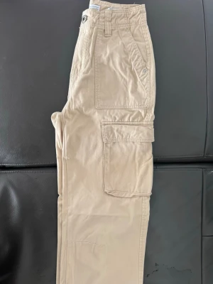 Bershka Beiga cargobyxor med stora fickor - Snygga beiga cargobyxor med raka ben och stora fickor på sidorna. Byxorna har klassisk cargomodell med flera praktiska fickor och stängs med knapp och dragkedja. Perfekta för en avslappnad streetstil. Materialet är bomull för en skön känsla.