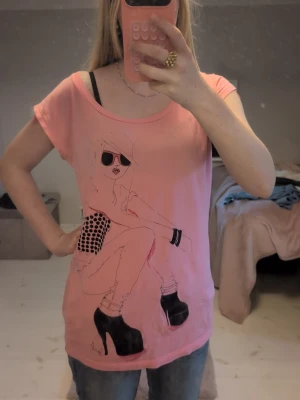 Rosa t-shirt med coolt tryck - Rosa t-shirt med ett stort tryck av en tecknad tjej i solglasögon och platåskor. T-shirten har rund halsringning och korta ärmar. Materialet känns mjukt och skönt, troligen bomull eller polyester. Perfekt för dig som gillar statement-plagg. Ganska stor tröja är mer som en medium.