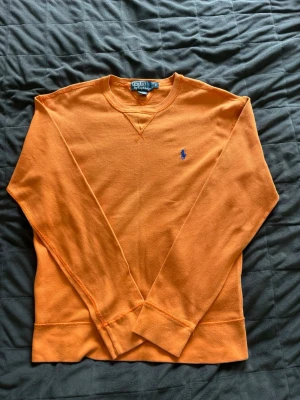 Orange stickad tröja från Polo Ralph Lauren - Snygg orange stickad tröja från Polo Ralph Lauren med rund halsringning och blå broderad logga på bröstet. Tröjan har långa ärmar och ribbade muddar vid ärmslut och nederkant. Perfekt för dig som gillar färg och klassisk stil.