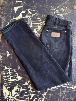 Svarta Wrangler straight leg jeans - Säljer ett par svarta jeans från Wrangler med klassisk straight leg passform. Jeansen har fem fickor, bälteshällor och detaljer i kopparfärg. Materialet är kraftig denim i bomull och bakfickan har Wrangler-lappen i läder.