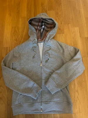 Grå hoodie från Burberry med dragkedja - Säljer en grå hoodie från Burberry med klassiskt rutigt foder i huvan och broderad logga på bröstet. Hoodien har snörning i huvan, dragkedja framtill och ribbade muddar. Perfekt för dig som vill ha en stilren och lyxig look.