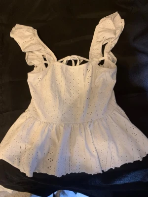 Vit broderad linnetopp med volang - Supersöt vit linnetopp med broderade detaljer och volangkant. Toppen har breda axelband med volanger och öppen rygg med knytband. Perfekt för varma dagar. 