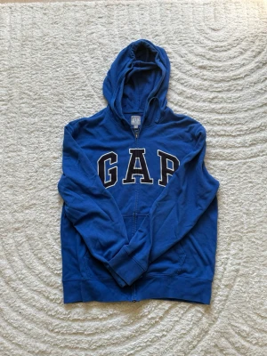 Blå hoodie med dragkedja från GAP - Snygg blå hoodie från GAP med dragkedja framtill och stor vit logga på bröstet. Hoodien har huva, känguruficka och ribbade muddar vid ärmslut och nederkant. Perfekt för en avslappnad och sportig stil.