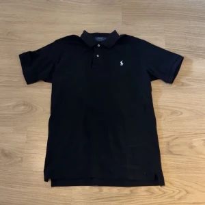 Svart pikétröja från Polo Ralph Lauren - Klassisk svart pikétröja från Polo Ralph Lauren med vit broderad logga på bröstet. Tröjan har krage, knappar framtill och korta ärmar. Perfekt för en stilren och avslappnad look.