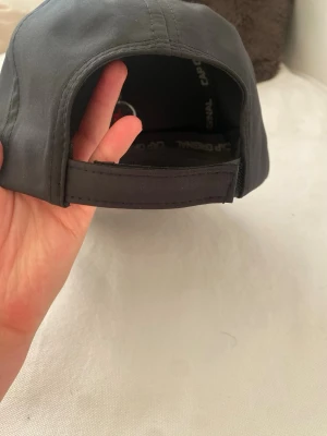 Mörkgrå keps från Cap Original - Snygg mörkgrå keps från Cap Original med klassisk böjd skärm och justerbart kardborreband bak. Kepsen är tillverkad i ett mjukt tygmaterial och har svarta detaljer på insidan med loggan tryckt på banden.