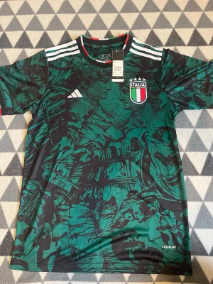 Italien matchtröja Adidas grön M - Snygg grön matchtröja från Adidas för Italiens landslag. Tröjan har ett unikt konstnärligt mönster i svart, Italiens klubbmärke på bröstet och vita Adidas-ränder på axlarna. Rund hals med italienska flaggans färger och tillverkad i lätt funktionsmaterial.