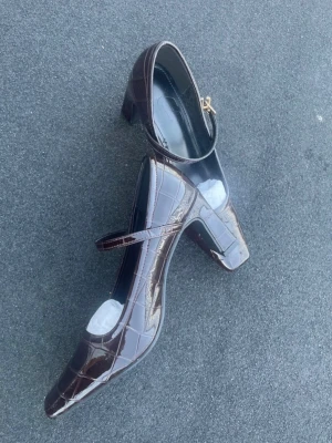 Bruna lackade Mary Jane-pumps - Säljer ett par mörkbruna Mary Jane-pumps i glansigt lackmaterial med krokodilpräglat mönster. Skorna har fyrkantig tå, bred klack och justerbar rem med guldfärgat spänne. Perfekta för dig som gillar en retrovibe och vill sticka ut.