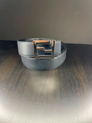 Svart bälte med metallspänne - Snyggt svart bälte med ett stort, fyrkantigt metallspänne med unik design. Bältet har en stilren look och passar perfekt till jeans eller kostymbyxor. Tillverkat i ett slätt material som ger en exklusiv känsla.