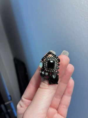 Svart statementring med stenar - Unik svart statementring med flera olika stenar i svart, vitt och genomskinligt. Ringen har en stor svart fyrkantig sten i mitten, omgiven av mindre gnistrande stenar och en vit opalliknande detalj. Designen är modern och edgy med flera lager och detaljer. Lite avskavt på ena sidan som ni ser på bild. 