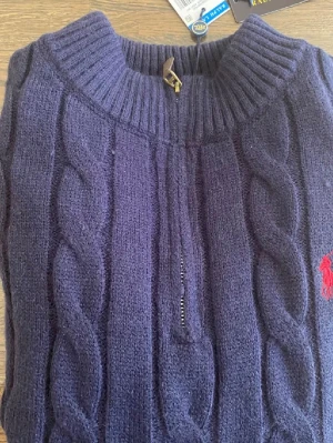 Mörkblå kabelstickad tröja Polo Ralph Lauren - Kabelstickad mörkblå tröja från Polo Ralph Lauren med halv dragkedja och ribbad hög krage. Tröjan har det klassiska röda Polo-logot broderat på bröstet och är gjord i ett mjukt, tjockt material. Perfekt för kalla dagar och snygg till jeans.