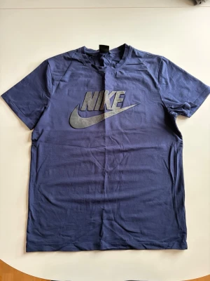 Blå Nike t-shirt med logga - Snygg blå t-shirt från Nike med stor silverfärgad logga på bröstet. Klassisk rak passform och korta ärmar. Tillverkad i mjukt bomullsmaterial som känns skönt mot huden. Perfekt för en sportig och avslappnad stil.