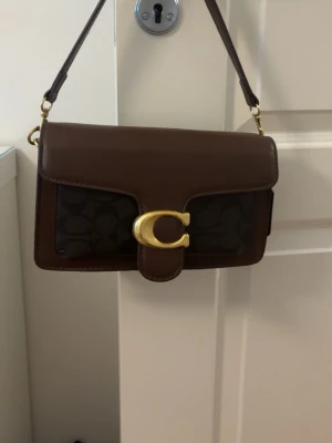 Coach väska - Snygg brun handväska från Coach i läder med klassiskt monogrammönster och guldfärgat C-spänne framtill. Väskan har en kort axelrem och stilren, rektangulär form. Perfekt för dig som vill ha en trendig och tidlös accessoar.