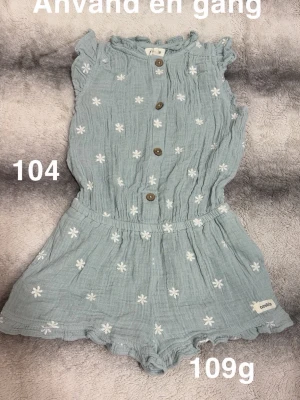 Blå jumpsuit med blommor och knappar - Supersöt blå jumpsuit med vita broderade blommor och träknappar framtill. Ärmlös modell med volangkant vid axlar och ben, samt resår i midjan för skön passform. Tillverkad i mjukt, luftigt tyg som passar perfekt till sommaren.🌸 Använd en gång. Storlek 104
