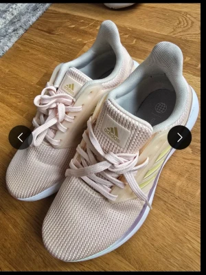 Ljusrosa Adidas sneakers med mesh - Snygga ljusrosa Adidas sneakers med mesh-ovandel och klassiska tre ränder i ljusgul på sidan. Skorna har vita sulor med en touch av lila och beige detaljer. Lätta och bekväma med snörning och vadderad insida. Perfekta för en sportig och trendig look.