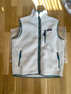 Vit fleeceväst från Patagonia - Mysig vit fleeceväst från Patagonia med hög krage och dragkedja framtill. Västen har tre fickor med dragkedja och gröna detaljer längs kanterna och dragkedjorna. Perfekt för lager-på-lager och outdoor-vibes.