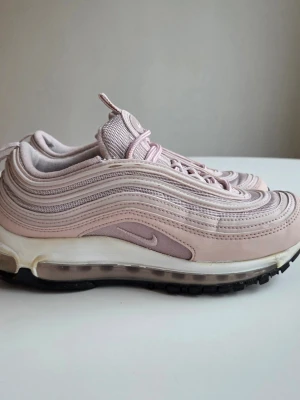 Nike Air Max 97 rosa sneakers - Säljer ett par Nike Air Max 97 i en snygg ljusrosa färg. Skorna har den ikoniska vågiga designen, synlig Air-sula och detaljer i mesh och syntetmaterial. Perfekta för dig som vill ha en clean och sportig look med streetkänsla. Obs bilderna för detaljer.