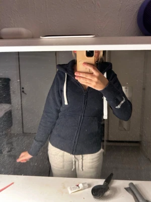 Mörkblå hoodie från russemerch  - Mörkblå zip-hoodie från russemerch med vit dragsko i huvan och broderad logga på ärmen. Hoodien har fickor framtill och ribbade muddar vid ärmslut och nederkant. Perfekt för en avslappnad och sportig stil.