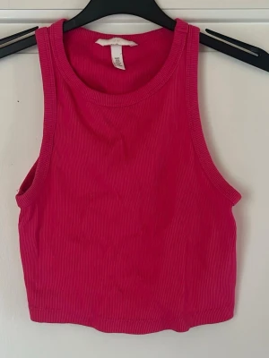 Rosa ribbad crop top från H&M - Säljer en snygg rosa ribbad crop top från H&M. Toppen har breda axelband, rund halsringning och är tillverkad i ett stretchigt material som sitter tajt och bekvämt. Perfekt till jeans eller kjol för en trendig look.