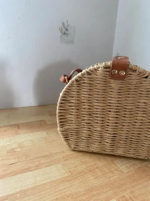 Helt ny rund flätad stråväska - Säljer en rund handväska i naturfärgat flätat stråmaterial med bruna läderdetaljer och axelrem. Väskan har ett lock som stängs med två guldfärgade knappar. Perfekt för strand och sol. Helt ny handväska 