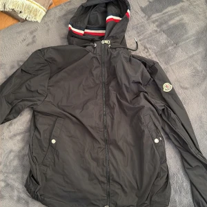 Svart vindjacka från Moncler med huva - Svart vindjacka från Moncler med huva som har snygga ränder i rött, vitt och blått. Jackan har dragkedja framtill, två fickor med tryckknappar och Moncler-logga på ärmen. Perfekt för dig som gillar stilrena och sportiga plagg. Passar liten xl eller stor L