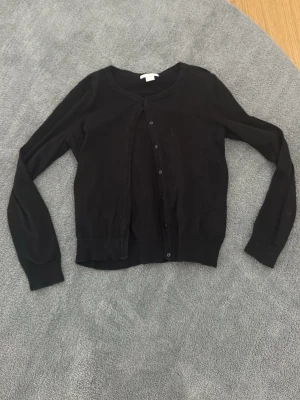 Svart basic kofta från H&M - En enkel svart kofta från H&M Basic med rund halsringning och knäppning framtill. Långärmad modell i mjukt och tunt material, perfekt att slänga över en topp eller klänning. Klassisk och stilren design som funkar till det mesta.