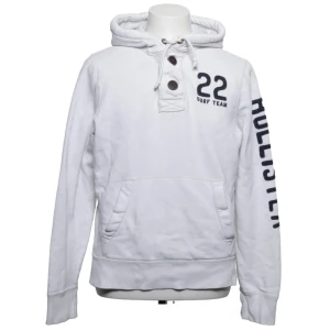 Vit hoodie från Hollister med tryck - Vit hoodie från Hollister med svart tryck på ena ärmen och siffran 22 samt 'Surf Team' på bröstet. Hoodien har knappar vid halsen, dragsko i huvan och en stor magficka. Tillverkad i mjukt bomullsmaterial och har ribbade muddar vid ärmslut och nederkant.