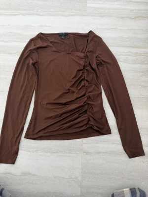 Brun långärmad topp från Amisu - Snygg brun långärmad topp från Amisu med rynkade detaljer längs ena sidan framtill. Tillverkad i ett stretchigt och mjukt material som sitter tajt och bekvämt. Perfekt för dig som gillar en enkel men trendig look.