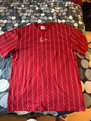 Karl Kani Oversized T - Säljer en röd oversized t-shirt från Karl Kani med vita vågiga ränder över hela tröjan och en broderad logga på bröstet. T-shirten har rund halsringning och långa ärmar. Perfekt för dig som gillar streetwear och vill sticka ut lite extra.