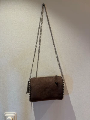 Brun axelväska i mocka med kedja - Snygg brun axelväska i mocka med silverfärgade kuldetaljer längs kanterna och en lång kedjerem. Väskan har en minimalistisk design och stängs med dragkedja. Perfekt för dig som gillar en stilren och trendig look.