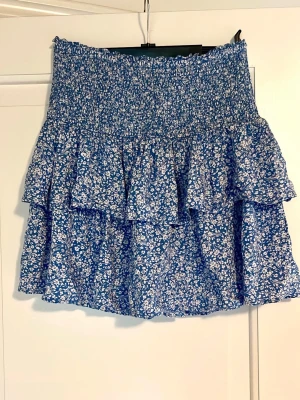 Blommig blå volangkjol från Kappahl - Supersöt blå kortkjol från Kappahl med vitt blommönster och volanger. Kjolen har smock i midjan för skön passform och är perfekt för vår och sommar. Lätt och luftig, passar till både sneakers och sandaler.