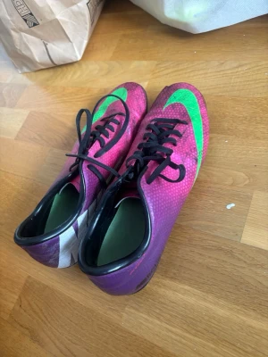 Nike Mercurial lila/gröna fotbollsskor - Säljer ett par Nike Mercurial fotbollsskor i lila med gröna detaljer och svart sula. Skorna har snörning och är designade för spel på gräs. Ovandelen är tillverkad i syntetmaterial för bra bollkontroll och lätt vikt. Perfekta för dig som vill sticka ut på planen.