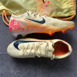 Nike Mercurial Elite fotbollsskor  - Säljer ett par Nike Mercurial fotbollsskor i ljusgul med svarta och neonrosa detaljer. Skorna har snörning, svart Nike-logga och mönstrad sula i orange och rosa. Det är även mbappes egna design på skorna då det är hans egna sko så att säga, i storlek 47,5