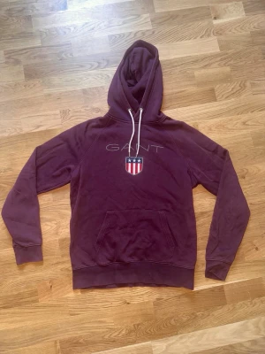 Vinröd hoodie från GANT med logga - Säljer en vinröd hoodie från GANT med vit dragsko i huvan och stor GANT-logga samt sköld med stjärnor och ränder på bröstet. Hoodien har känguruficka framtill och ribbade muddar vid ärmslut och nederkant. Perfekt för en chill och snygg look.