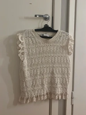Zara topp - Supersöt vit stickad topp från Zara med volangdetaljer vid ärmslut och nederkant. Toppen har ett fint spetsliknande mönster och är ärmlös, perfekt för en fräsch och trendig look.