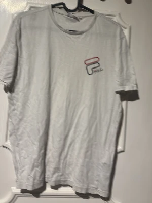 Vit t-shirt från Fila med logga - Vit t-shirt från Fila med liten logga i rött, blått och grått på bröstet. Klassisk rund hals och korta ärmar. Perfekt för en avslappnad och sportig stil. Tillverkad i mjuk bomull för skön känsla hela dagen.