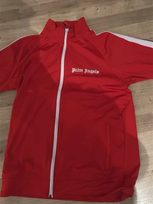 Röd zip-tröja från Palm Angels - Snygg röd zip-tröja från Palm Angels med vit dragkedja och vita detaljer på ärmarna. Tröjan har hög krage, två fickor och logga broderad på bröstet. Tillverkad i 90% polyester och 10% elastan för en sportig och bekväm känsla.
