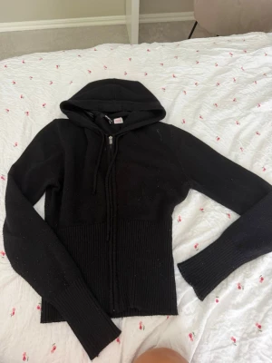 Svart hoodie från Gina Tricot - Svart hoodie från Gina Tricot med dragkedja framtill och ribbade muddar samt nederkant. Modellen har huva med dragsko och är tillverkad i ett mjukt stickat material. Perfekt för en chill och stilren look.