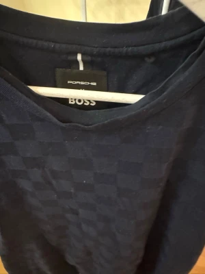 Mörkblå rutig t-shirt Porsche BOSS - Snygg mörkblå t-shirt från Porsche BOSS med diskret rutigt mönster. Klassisk rund halsringning och korta ärmar. Perfekt för dig som gillar stilrena plagg med en twist. Tillverkad i mjuk bomull för skön känsla hela dagen.