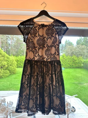 Svart spetsklänning med kort ärm - Kort klänning med beige underklänning och svart spets ovanpå. Klänningen har rund hals, korta ärmar och ett detaljerat spetsmönster med blommor och cirklar. Passformen är normal och kjolen har lätt volym. Perfekt för dig som gillar romantisk stil.
