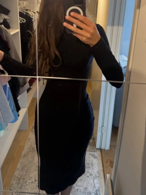 Svart långärmad midiklänning - Snygg svart klänning med lång ärm och figurnära passform. Klänningen har en slits på sidan och hög hals, vilket ger en stilren och modern look. Perfekt för dig som gillar minimalistisk stil och vill ha något enkelt men ändå elegant.