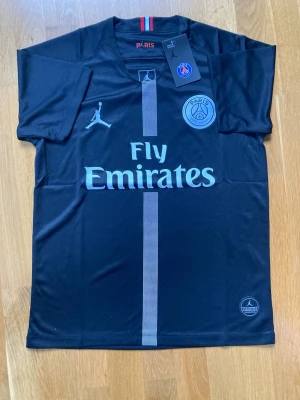 Svart PSG matchtröja Jordan x Fly Emirates - Snygg svart PSG matchtröja i samarbete med Jordan och Fly Emirates. Tröjan har korta ärmar, rund hals och PSG-logga på bröstet. Tillverkad i lätt och ventilerande funktionsmaterial, perfekt för fotboll eller träning.