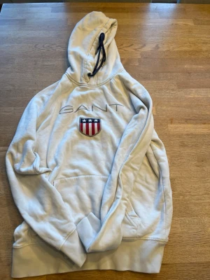 Vit hoodie från GANT med logga - Säljer en vit hoodie från GANT med broderad logga och ett sköldmärke med stjärnor och ränder på bröstet. Hoodien har huva med svart dragsko och en stor magficka. Perfekt för dig som gillar klassisk college-stil. Storlek xs