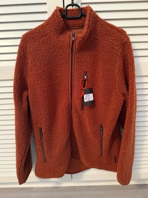  Hansen & Jacob fleece helt ny - Snygg rostbrun teddyjacka från Hansen & Jacob med hög krage och dragkedja framtill. Jackan har två praktiska fickor med dragkedja och är tillverkad i mjukt, fluffigt teddy-material som håller dig varm. Perfekt för dig som gillar en avslappnad och trendig stil helt ny 900 kr 