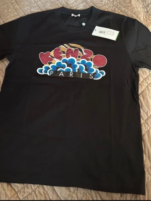 Svart Kenzo t-shirt med tryck - Säljer en svart t-shirt från Kenzo med färgglatt tryck på bröstet där det står 'Kenzo Paris' i rött, blått och vitt. T-shirten har klassisk passform och rund hals. Perfekt för dig som vill ha en statement tee med snygg design.