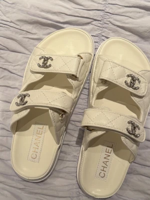 Vita sandaler  - Säljer ett par vita sandaler med två breda remmar och silverfärgade CC-loggor. Sandalerna har en chunky sula och kardborreknäppning för enkel justering. Perfekta för en trendig och lyxig look. Storlek 38!Aldrig använda 💕