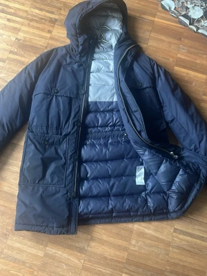 Mörkblå parkas från J.Lindeberg - Snygg mörkblå parkas från J.Lindeberg med huva och dragkedja. Jackan har flera praktiska fickor framtill och quiltad insida för extra värme. Perfekt för kyliga dagar och har en stilren, modern look.