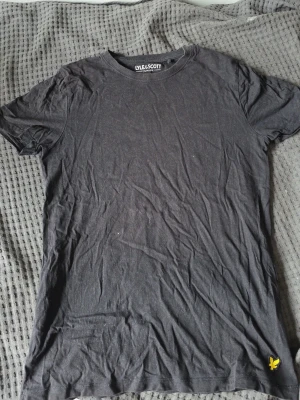 Svart t-shirt från Lyle & Scott - En klassisk svart t-shirt från Lyle & Scott med rund hals och korta ärmar. T-shirten har den ikoniska gula loggan broderad längst ner på framsidan. Tillverkad i mjuk bomull för en skön känsla och enkel stil. Passar xs
