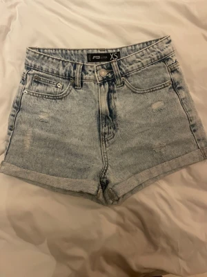 Ljusblå denimshorts från F3 Studio - Snygga ljusblå denimshorts från F3 Studio i klassisk femficksmodell. Shortsen har hög midja, uppvikta benslut och slitna detaljer framtill och baktill för en trendig look. Perfekta till sommaren och lätta att matcha med olika stilar.